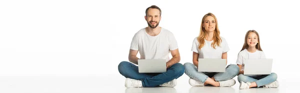 Fröhliche Familie auf dem Boden sitzend mit Laptops auf überkreuzten Beinen, isoliert auf weißen, panoramischen Aufnahmen — Stockfoto