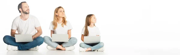 Family sitting on floor with laptops on crossed legs and looking away isolated on white, panoramic shot — Stock Photo