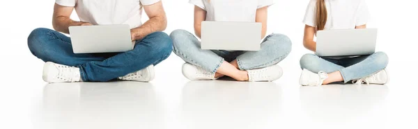 Cropped view of family sitting on floor with laptops on crossed legs on white, panoramic shot — Stock Photo