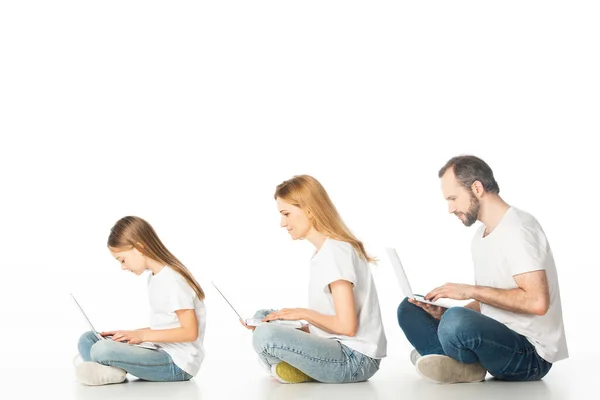 Vista lateral de la familia sentada en el suelo con computadoras portátiles en piernas cruzadas aisladas en blanco - foto de stock