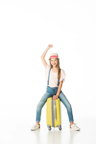 Fröhliches Mädchen mit Hut sitzt auf gelber Reisetasche isoliert auf weißem Grund — Stockfoto