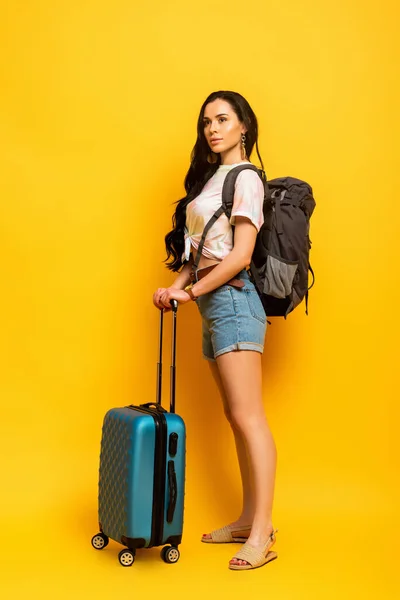 Brünette Frau mit Rucksack und Koffer auf gelbem Hintergrund — Stockfoto