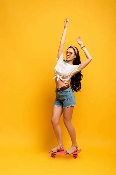 Stylish summer brunette girl riding pink penny board with hands in air on yellow background — Stock Photo