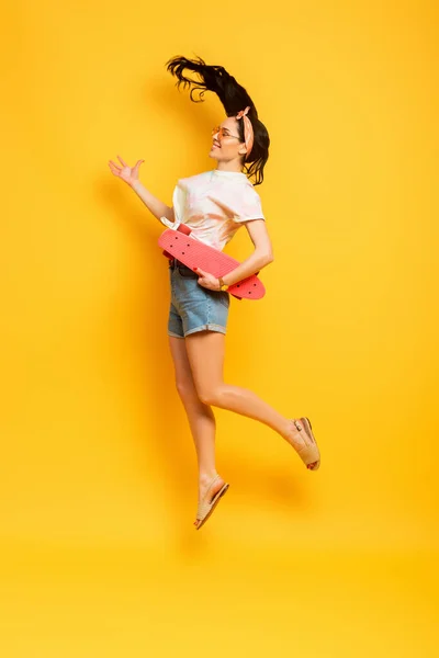 Vista lateral de la feliz chica morena de verano elegante saltando con tablero de penique rosa sobre fondo amarillo - foto de stock
