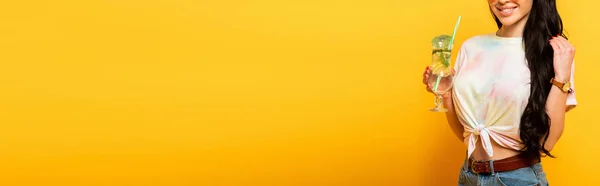 Обрезанный вид на улыбающуюся стильную летнюю брюнетку с освежающим коктейлем на желтом фоне, панорамный снимок — стоковое фото