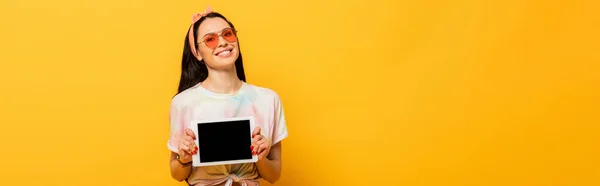 Усміхнена стильна літня брюнетка дівчина тримає цифровий планшет з порожнім екраном на жовтому тлі, панорамний знімок — стокове фото