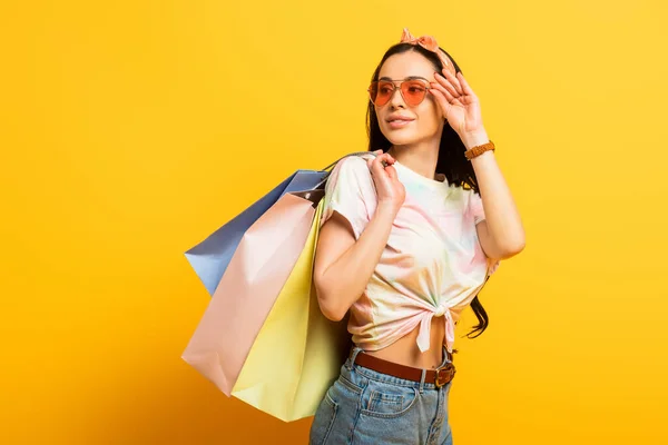 Sonriente elegante chica morena de verano con bolsas de compras mirando hacia otro lado sobre fondo amarillo - foto de stock