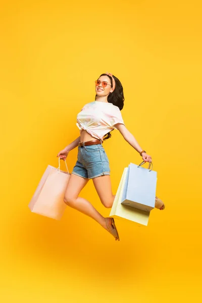 Full length view of smiling stylish summer brunette girl jumping with shopping bags on yellow background — Stock Photo
