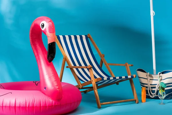 Sedia a sdraio a strisce vicino a fenicottero gonfiabile, crema solare, borsa da spiaggia e cocktail su sfondo blu — Foto stock