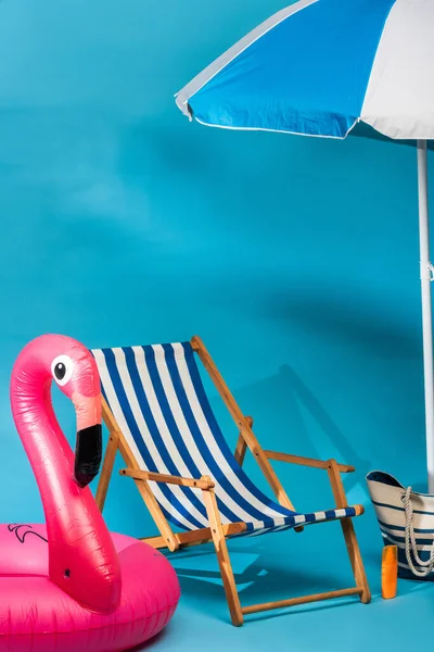 Silla de cubierta rayada cerca del flamenco inflable, protector solar, bolso de playa y paraguas sobre fondo azul - foto de stock