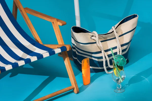 Vista da vicino della sedia a sdraio a strisce vicino alla protezione solare, borsa da spiaggia e cocktail su sfondo blu — Foto stock