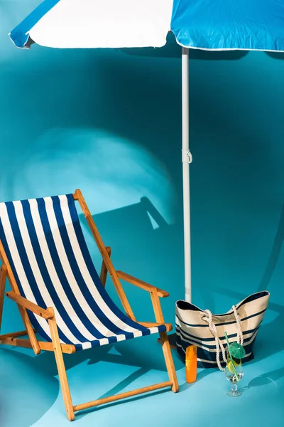Chaise longue rayée près de la crème solaire, sac de plage et cocktail sous parapluie sur fond bleu — Photo de stock