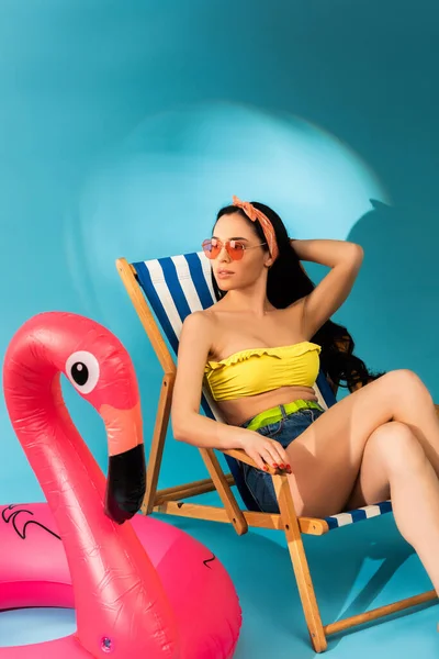 Stylish girl sitting in deck chair near inflatable flamingo on blue background — Stock Photo