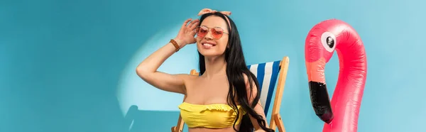 Fille élégante et souriante assise sur une chaise longue près d'un flamant rose gonflable sur fond bleu, plan panoramique — Photo de stock