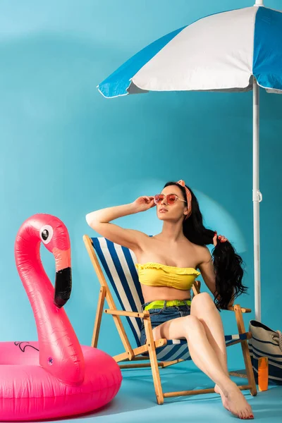 Chica elegante sentada en la silla de cubierta cerca de flamenco inflable, bolso de playa y paraguas sobre fondo azul - foto de stock