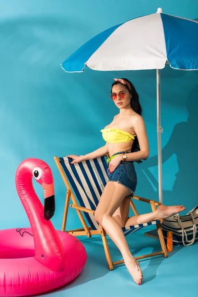 Élégante fille assise posant sur la chaise longue près de flamant rose gonflable, sac de plage et parapluie sur fond bleu — Photo de stock