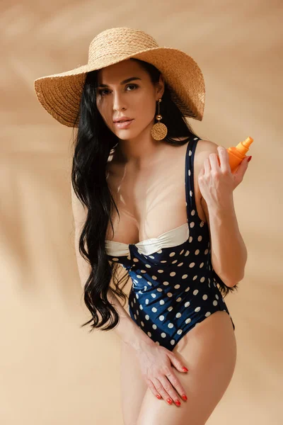 Sexy brunette woman in polka dot swimsuit and straw hat applying sunscreen on beige background — Stock Photo