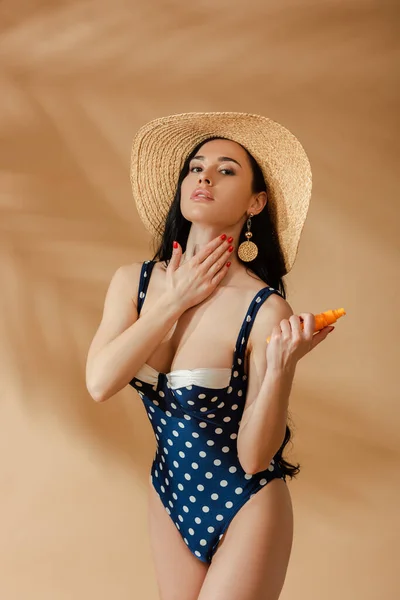Sexy brunette woman in polka dot swimsuit and straw hat applying sunscreen on beige background — Stock Photo