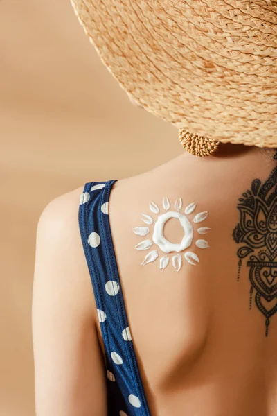 Woman in polka dot swimsuit and straw hat with drawn sun and tattoo on back on beige background — Stock Photo