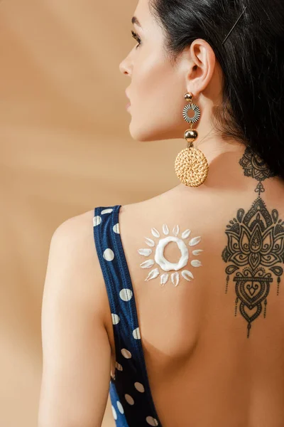 Back view of sexy brunette woman in polka dot swimsuit with drawn sun made of sunscreen and tattoo on back on beige background — Stock Photo