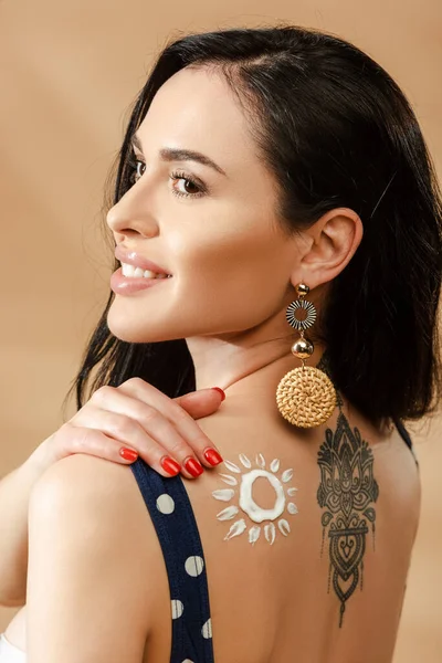 Smiling brunette woman in polka dot swimsuit with drawn sun made of sunscreen and tattoo on back on beige background — Stock Photo