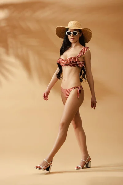 Fashionable sexy brunette woman in striped swimsuit, straw hat and sunglasses walking on beige background — Stock Photo