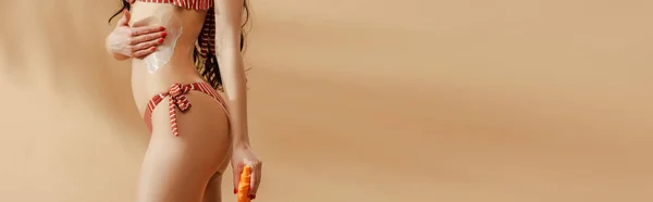 Обрізаний вид сексуальної брюнетки в смугастому купальнику, що наносить сонцезахисний крем на тіло на бежевому фоні, панорамний знімок — стокове фото