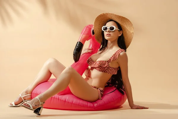 Sexy brunette woman in striped swimsuit, sunglasses and straw hat on inflatable flamingo on beige background — Stock Photo