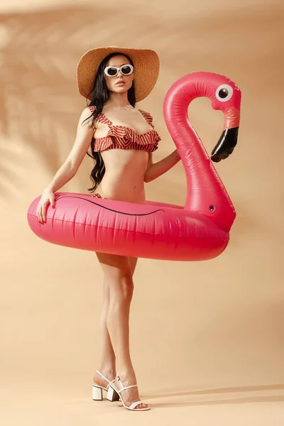 Sexy donna bruna in costume da bagno a righe, occhiali da sole e cappello di paglia con fenicottero gonfiabile su sfondo beige — Foto stock