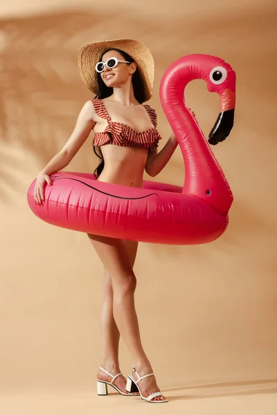 Femme brune sexy souriante en maillot de bain rayé, lunettes de soleil et chapeau de paille avec flamant rose gonflable sur fond beige — Photo de stock