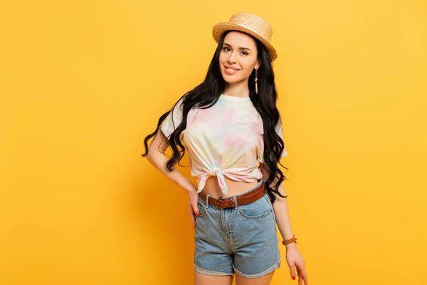 Smiling brunette girl in straw hat with hand on hip on yellow background — Stock Photo