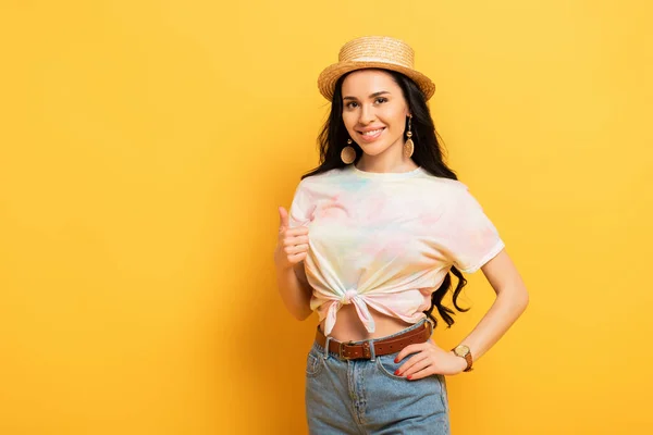 Smiling brunette girl in straw hat showing thumb up on yellow background — Stock Photo