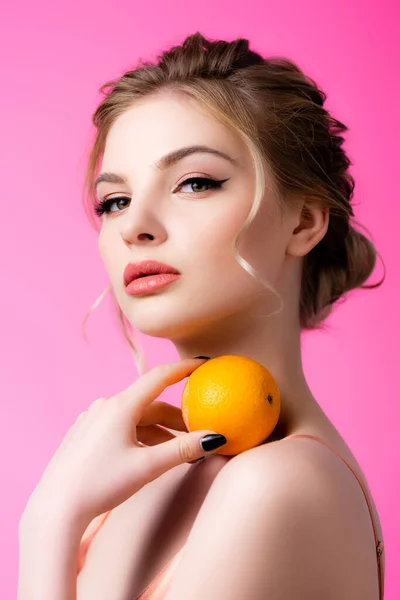 Elegante hermosa mujer rubia sosteniendo naranja madura en el hombro aislado en rosa - foto de stock