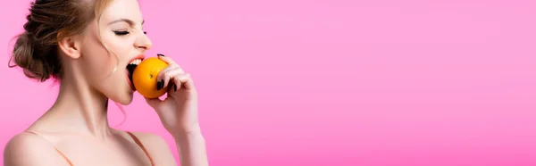 Seitenansicht der schönen blonden Frau beißt reife Orange isoliert auf rosa, Panoramaaufnahme — Stockfoto