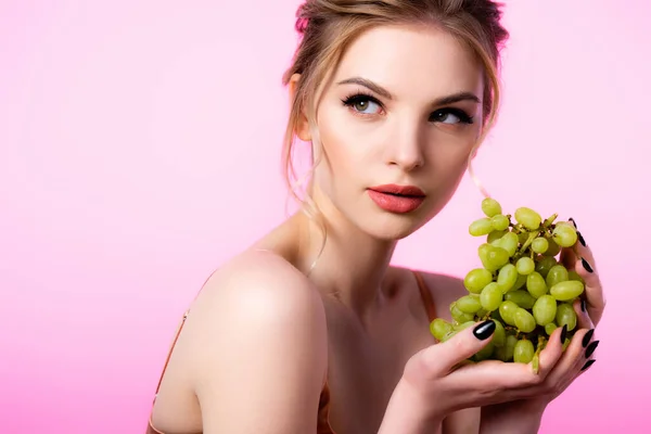 Elegante schöne blonde Frau hält grüne Weintrauben in der Hand und schaut isoliert auf rosa — Stockfoto