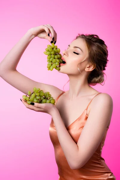 Elegante hermosa mujer rubia comiendo uvas verdes aisladas en rosa - foto de stock