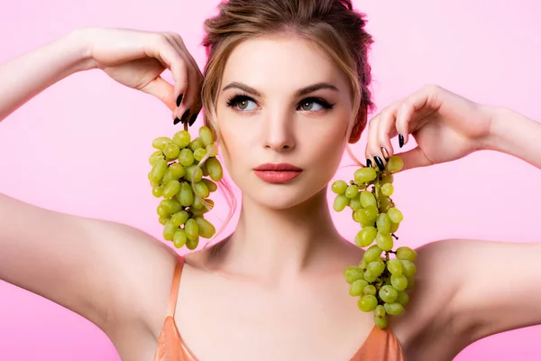 Елегантна красива блондинка тримає зелений виноград біля обличчя ізольовано на рожевому — стокове фото