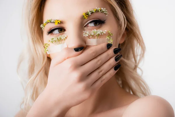 Naked beautiful blonde woman with wildflowers under eyes covering mouth isolated on white — Stock Photo