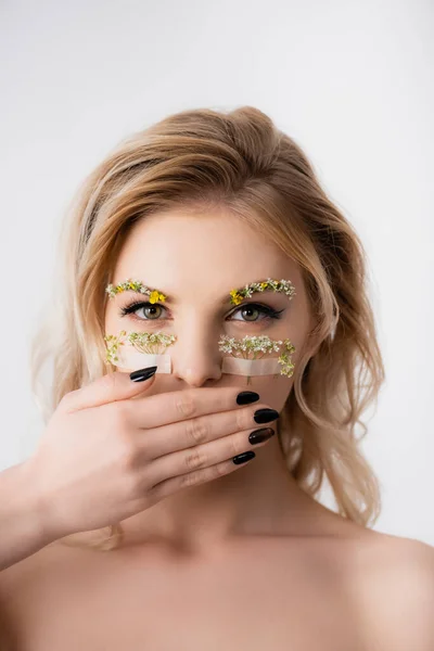 Naked beautiful blonde woman with wildflowers under eyes covering mouth isolated on white — Stock Photo