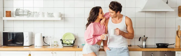 Panoramic crop of young woman embracing handsome boyfriend with cup of coffee in kitchen — Stock Photo