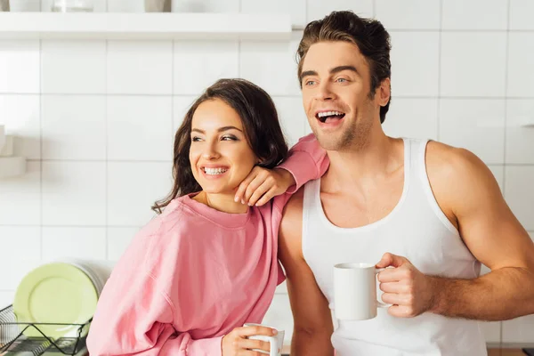 Donna positiva in possesso di una tazza di caffè e abbracciando bel fidanzato in cucina — Foto stock