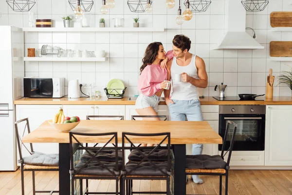 Focus selettivo della donna sexy che abbraccia il fidanzato con una tazza di caffè in cucina — Foto stock