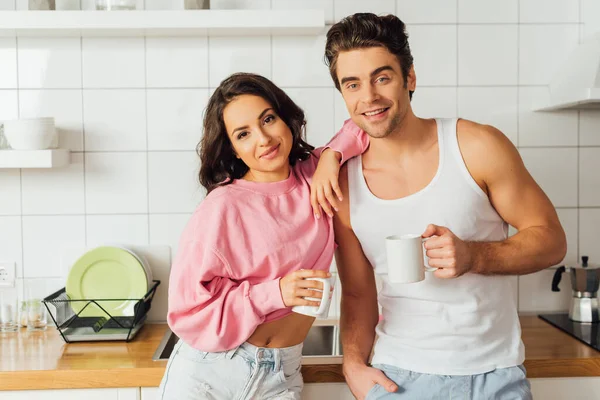 Sonriente pareja con tazas de café mirando a la cámara en la cocina - foto de stock