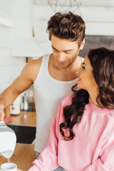 Sorrindo mulher olhando bonito namorado derramando água na xícara na cozinha — Fotografia de Stock
