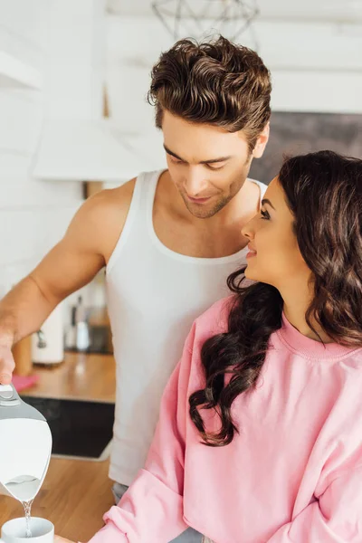 Femme souriante regardant beau petit ami verser de l'eau dans la tasse dans la cuisine — Photo de stock