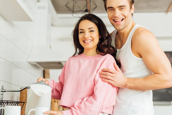 Красавчик обнимает весёлую девушку, держа чайник и чашку на кухне. — стоковое фото