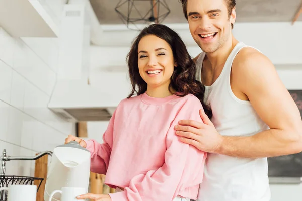Bonito homem abraçando alegre namorada segurando bule e xícara na cozinha — Fotografia de Stock