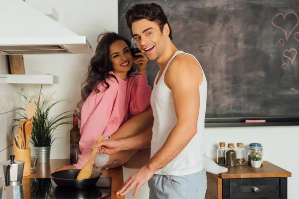 Enfoque selectivo del hombre sonriendo a la cámara mientras cocina cerca de su novia con un vaso de vino en la cocina - foto de stock