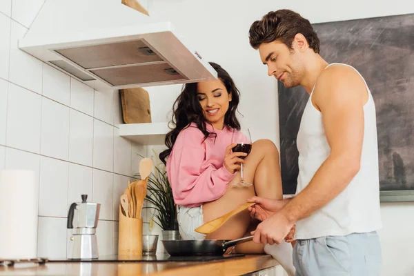 Selective focus of smiling girl holding glass of wine while sitting on worktop near boyfriend cooking in kitchen — Stock Photo