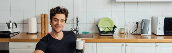 Panoramic shot of handsome man smiling at camera and holding cup of coffee in kitchen — Stock Photo